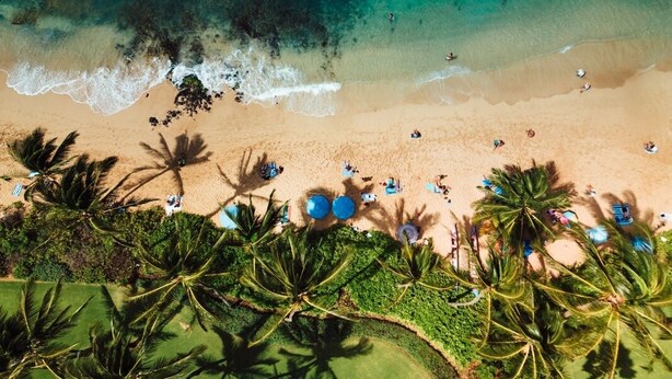 An aerial view of a beach with clear turquoise waters, palm trees, and people enjoying the beach and water activities. - Kauai Waikomo Stream Villas by Coldwell Banker Island Vacations (Koloa)