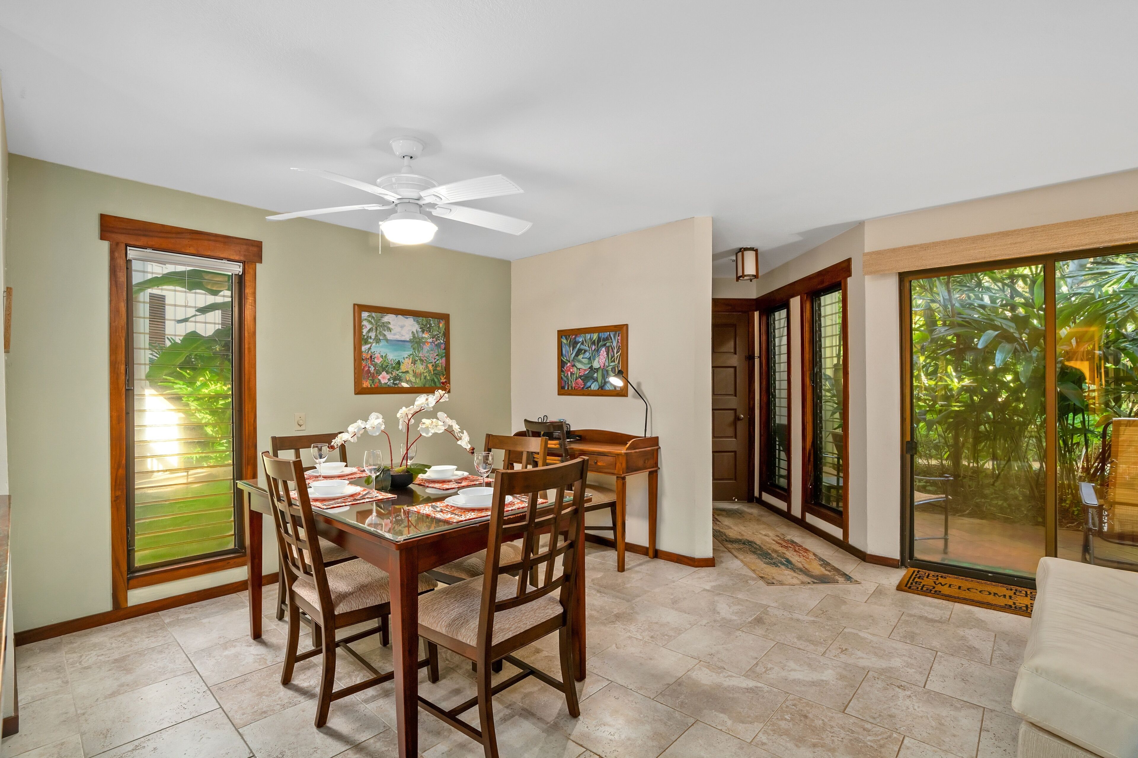 A dining area with a table set for four, wooden chairs, a ceiling fan, and a view through a glass door to a garden.