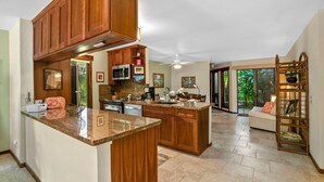 A spacious kitchen with wooden cabinets, granite countertops, and a large island. There is a dining area with a sofa and a window overlooking a lush garden. - Kauai Waikomo Stream Villas by Coldwell Banker Island Vacations (Koloa)