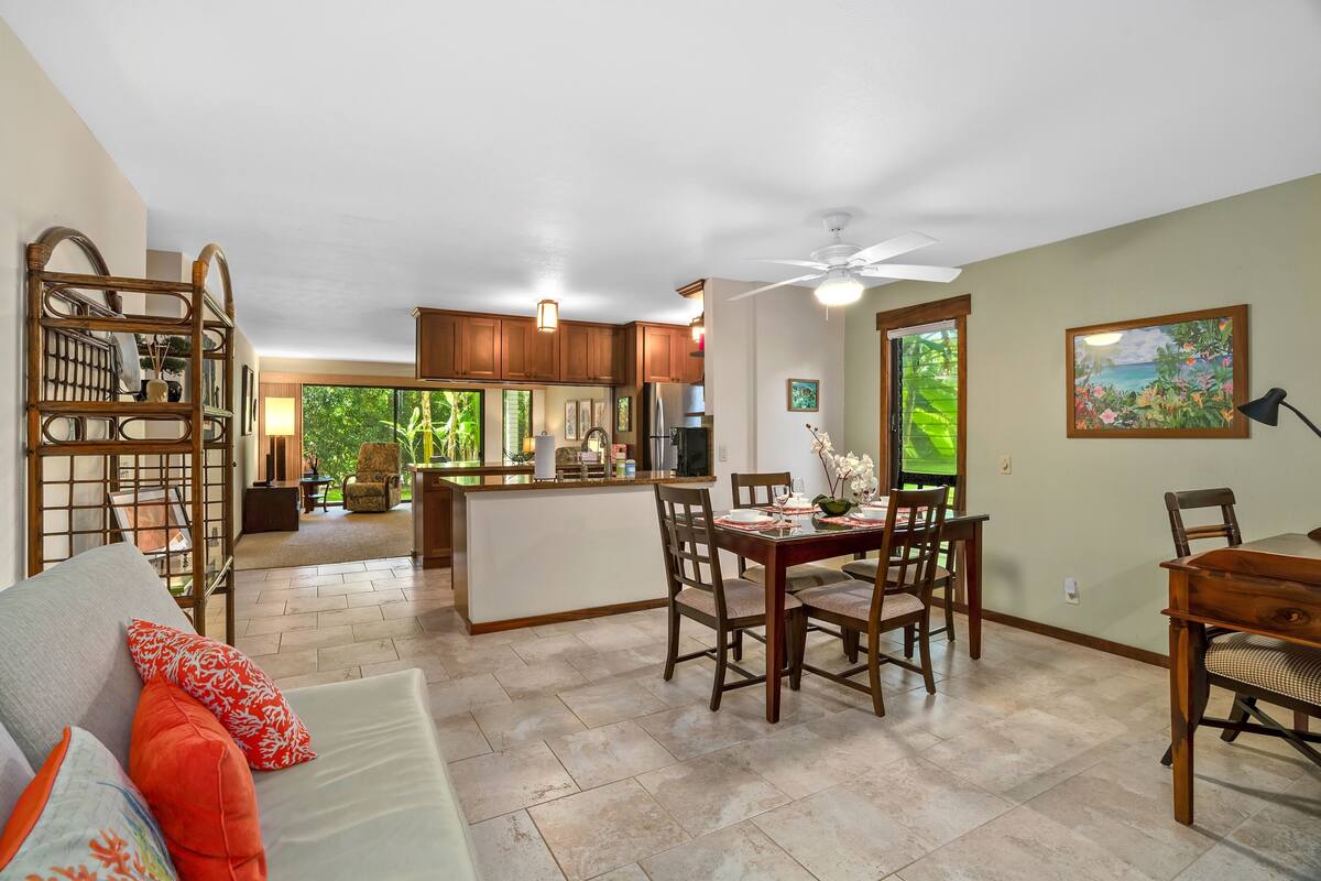 A spacious living area with a dining table, chairs, and a sofa with decorative pillows. A kitchen with wooden cabinets and a refrigerator is visible in the background.