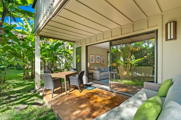 A covered patio with a table and chairs, a sofa, and a sliding glass door leading to a living room with a couch and a coffee table. - Kauai Waikomo Stream Villas by Coldwell Banker Island Vacations (Koloa)