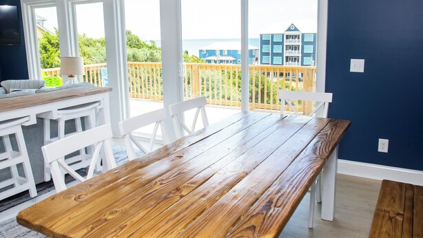 A wooden dining table with white chairs in a room with large windows. - Hot Mess - Pool, Oceanview, Pet-Friendly Home (Emerald Isle)