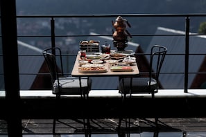 Enjoy a meal with a city view from this balcony dining area, featuring a table set with a plate of sliced meat, a bowl of salad, and a glass of tea.