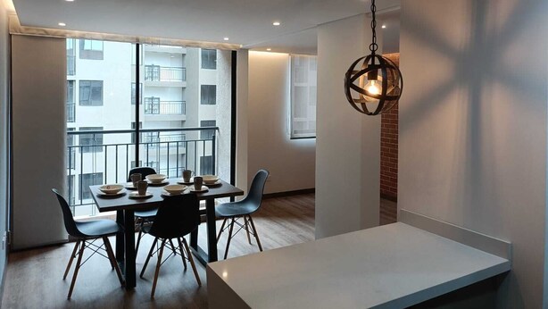 Un espace de repas moderne avec une table dressée pour quatre personnes, un luminaire suspendu et une vue sur un immeuble doté de balcons, visible à travers de grandes fenêtres.