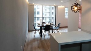 Un espace repas moderne avec un plancher en bois, une table blanche et des chaises bleues. Une grande fenêtre offre une vue sur les bâtiments à l’extérieur.