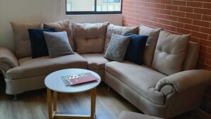 Un salon moderne avec un canapé d’angle beige, une table basse ronde et un mur d’accent en briques.