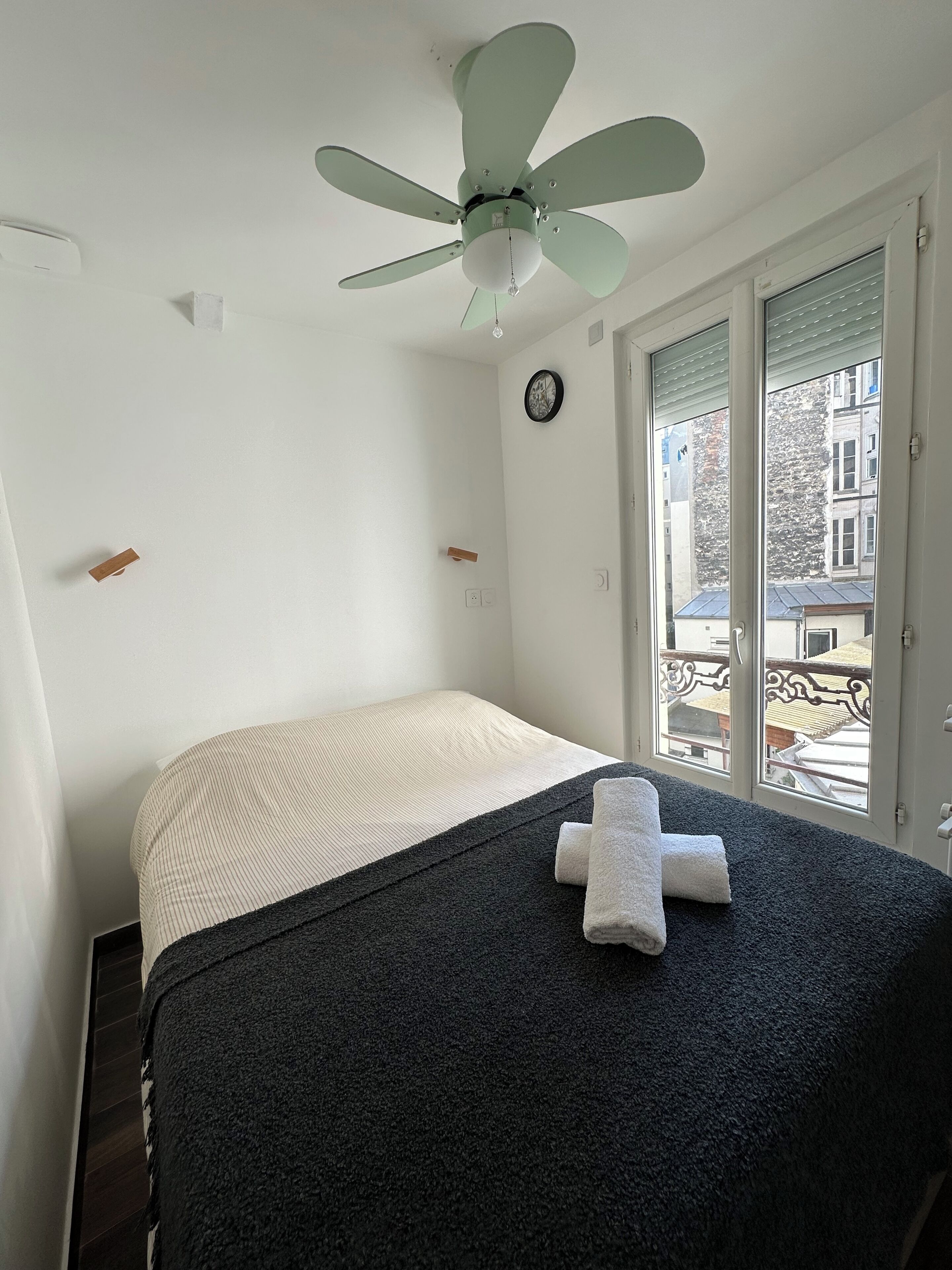 A bedroom with a bed, a ceiling fan, a clock, and a window with a view of buildings.