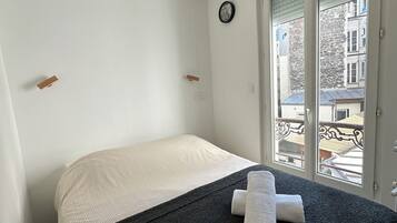A bedroom with a bed, a ceiling fan, a clock, and a window with a view of buildings.