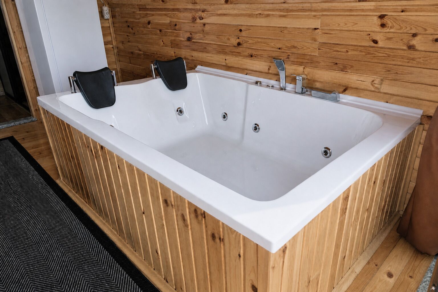 Jacuzzi rectangular con dos toallas negras, paneles de madera en las paredes y un tapete con estampado blanco y negro.