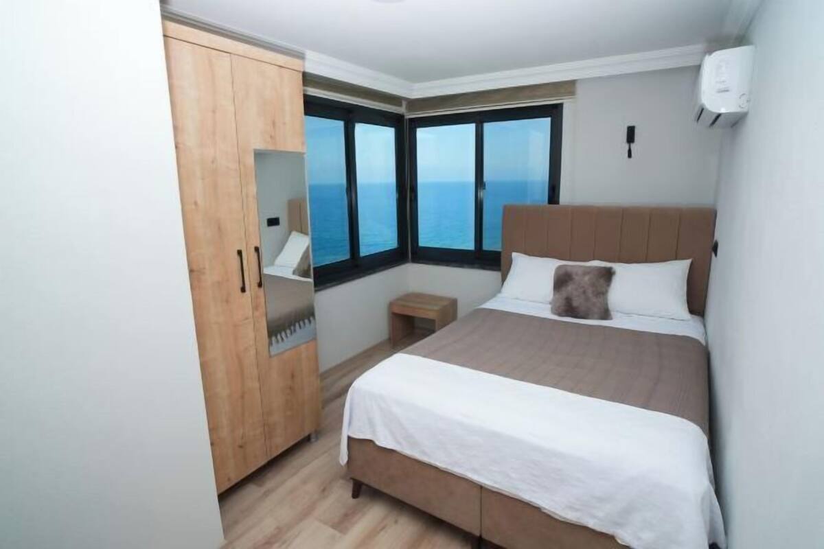 Une chambre à coucher avec un lit, une armoire en bois, une petite table de chevet et une vue sur la mer grâce à de grandes fenêtres.