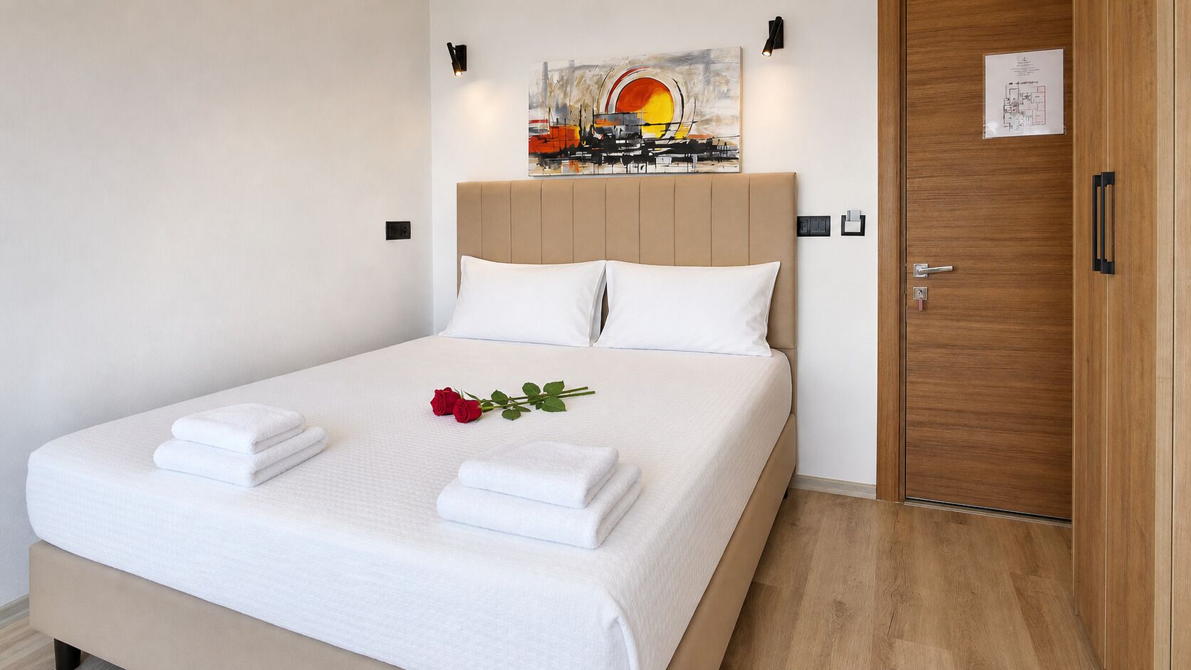 A neatly made bed with white linens, a single red rose, and two folded towels.