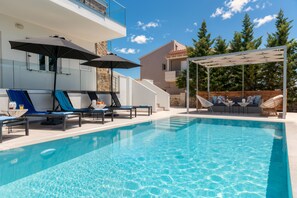 Ein Swimmingpool mit Liegestühlen und Sonnenschirmen, ein überdachter Sitzbereich und ein Gebäude mit Steinverkleidung.