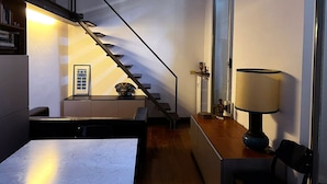 Une cuisine moderne avec un plan de travail en marbre, un coin repas avec une table et des chaises, un escalier avec une rampe en verre et une lampe sur une table d’appoint.