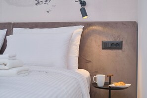 Ein ordentlich bezogenes Bett mit weißen Leinen, ein Nachttisch mit Tasse und Croissant sowie eine Wandleuchte.