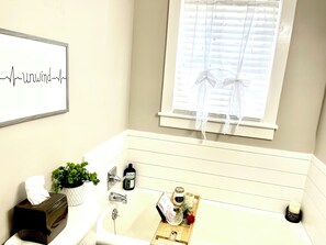 Ein Badezimmer mit einer weißen Badewanne, einem Fenster mit Jalousien, einem gerahmten Bild an der Wand, einer Packung Taschentücher, einer Topfpflanze und einer Kerze.