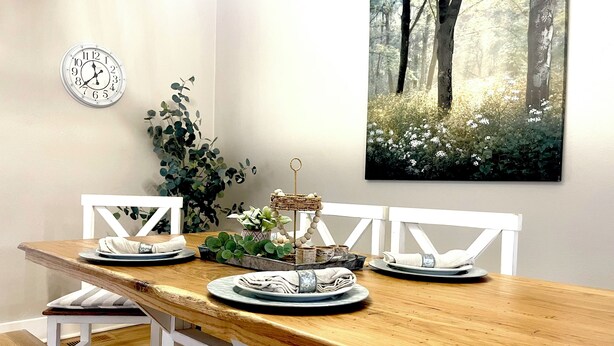 A dining area with a wooden table, white chairs, a clock, and a forest-themed painting on the wall. - “Delightful Decor” & “SO CLOSE” to Beech Bend, WKU, Caves, & Corvette Museum! (Bowling Green)