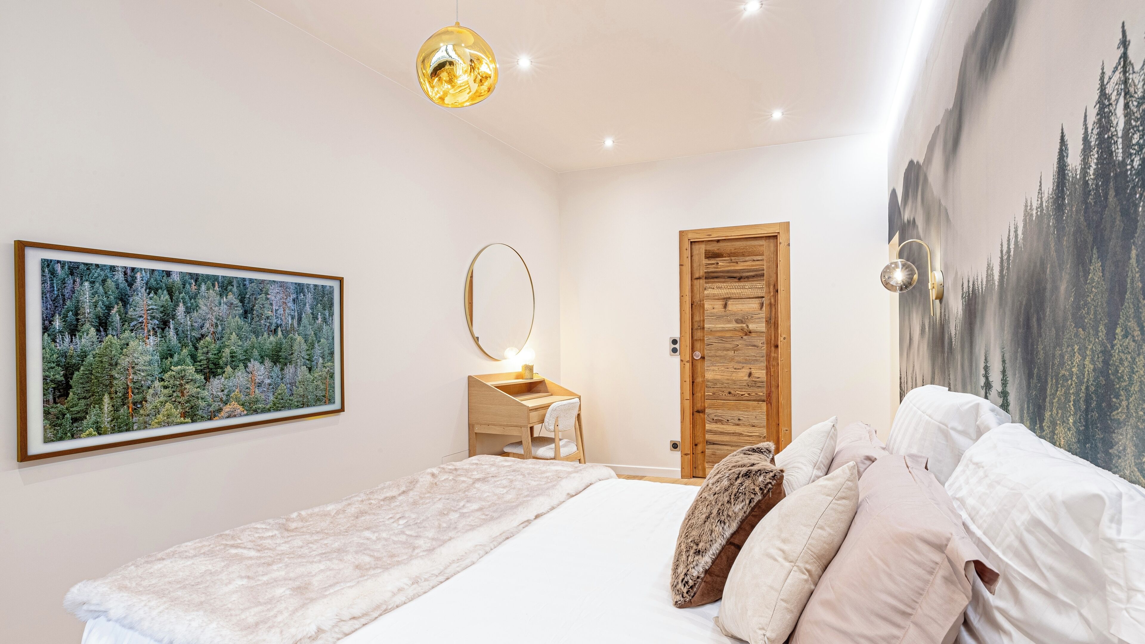 A bedroom with a bed, a desk, a mirror, and a large forest-themed painting.