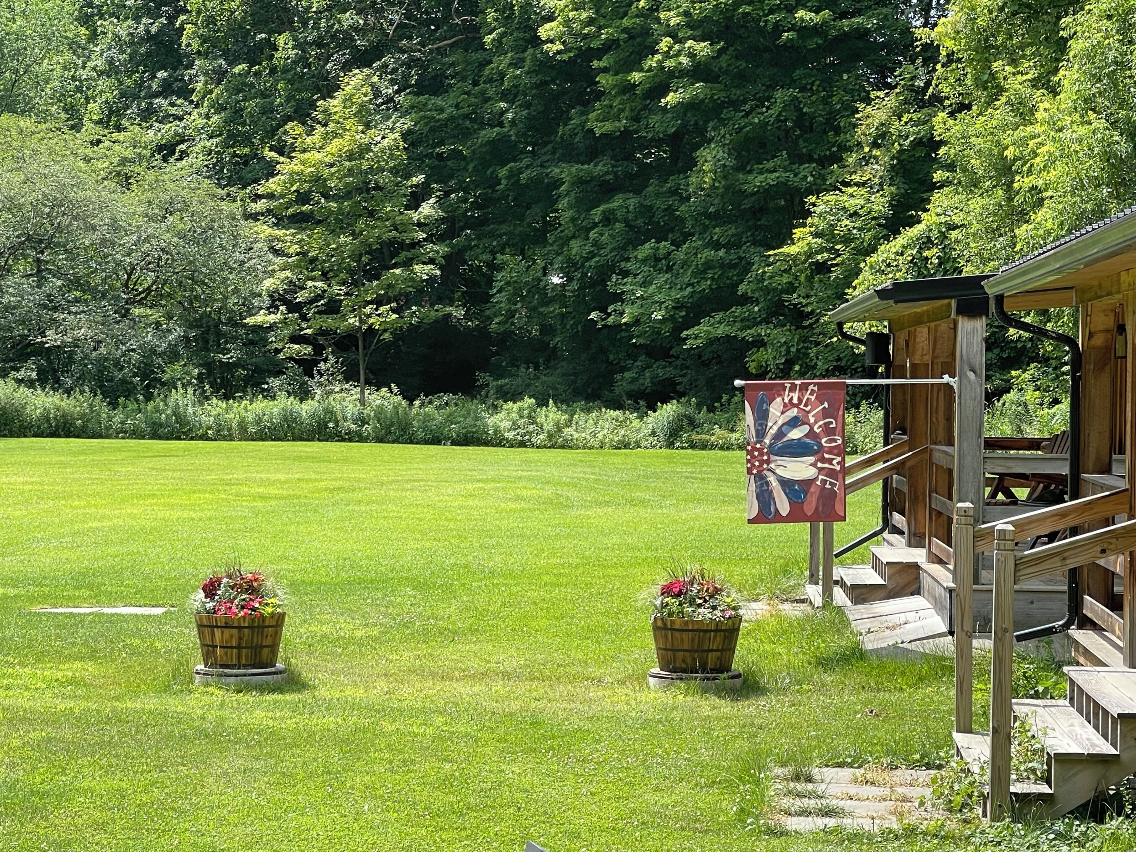 A wooden cabin with a 'WELCOME' sign, surrounded by a well-maintained lawn and two flower pots.