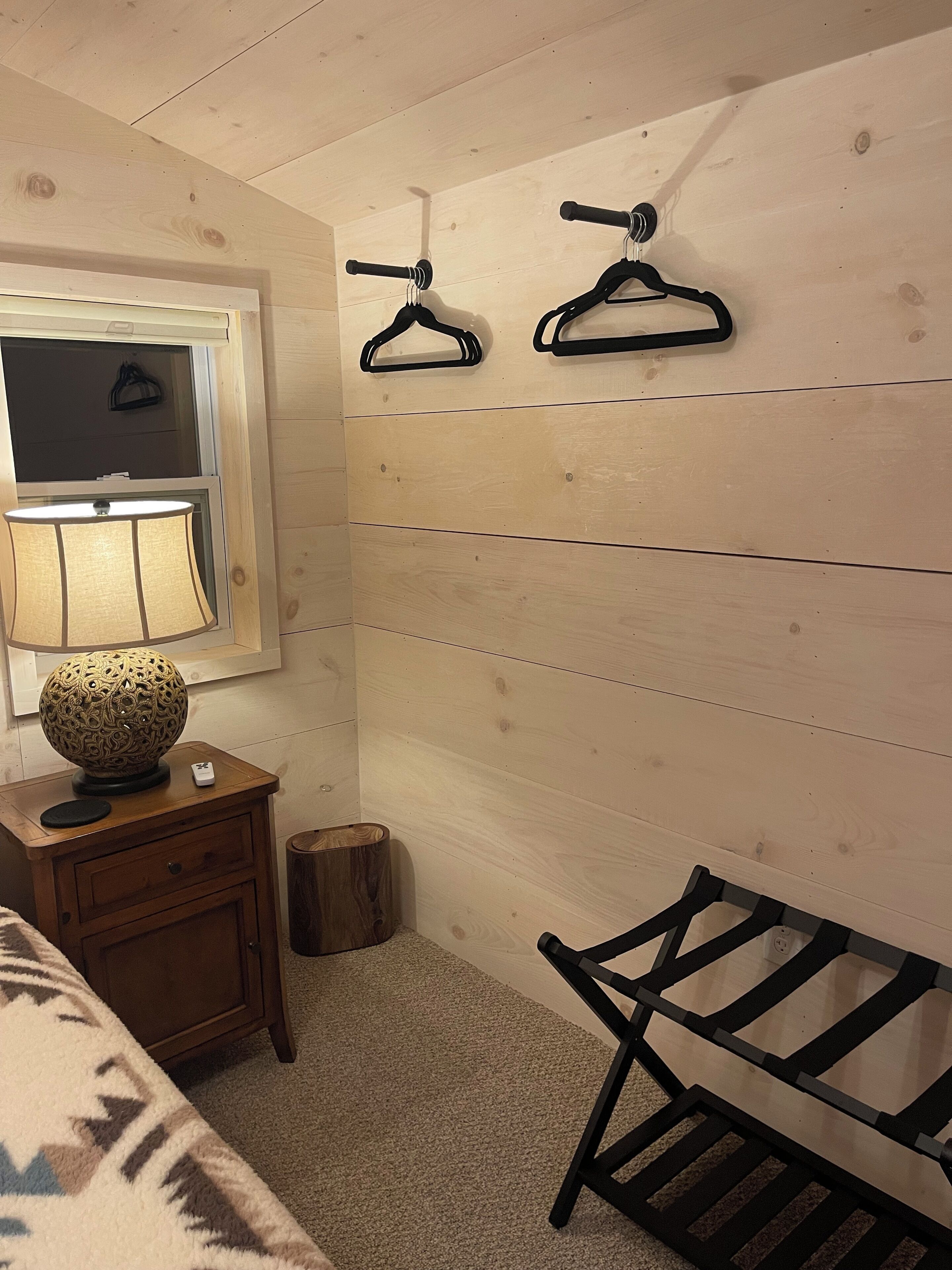 A room with a wooden floor, a bed with a patterned blanket, a bedside table with a lamp, a small wooden stool, and a chair with a metal frame.
