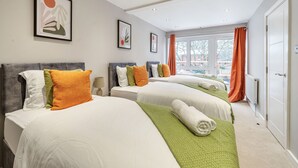 A hotel room with two beds, each with white bedding and colorful pillows, a green throw blanket, and a view of the outside through large windows with orange curtains. - Ettrick Court House By REDWOOD STAYS (Farnborough)
