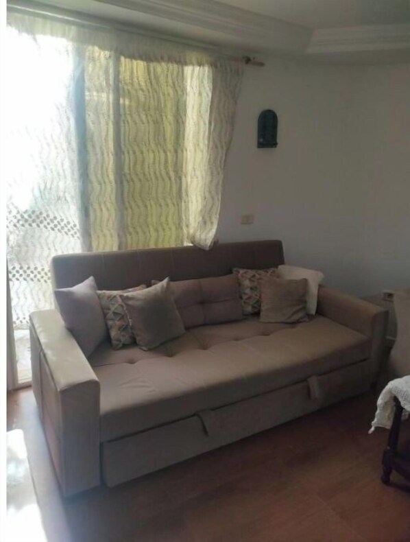 Ein beiges Sofa mit mehreren Kissen in einem Raum mit Holzboden und einem Fenster mit durchsichtigen Vorhängen.