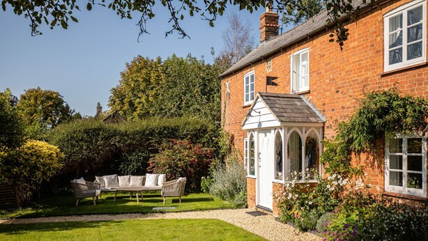 A brick house with a white door and window shutters, surrounded by a well-maintained garden with a lawn and various plants. - Stunning cottage with modern twists and original features - Carpenters Cottage (Sandford St Martin)