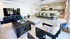 Ein modernes Wohnzimmer mit einem großen Flachbildfernseher, einem dunklen Holztisch und einer Chandelieber. Im Hintergrund befindet sich ein Essbereich mit einer Küche.