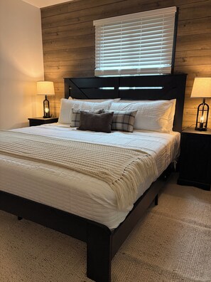Une chambre à coucher avec un lit, deux tables de chevet avec des lampes, une fenêtre avec des stores et une tête de lit en bois.