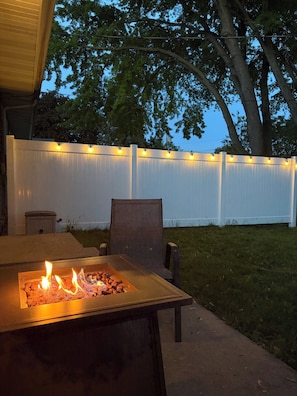Une terrasse extérieure avec une table à foyer en métal, une chaise et une clôture blanche décorée de guirlandes lumineuses.