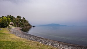 Un lac paisible, bordé d’une rive rocheuse, avec une zone herbeuse et une colline parsemée d’arbres et surmontée d’un bâtiment.