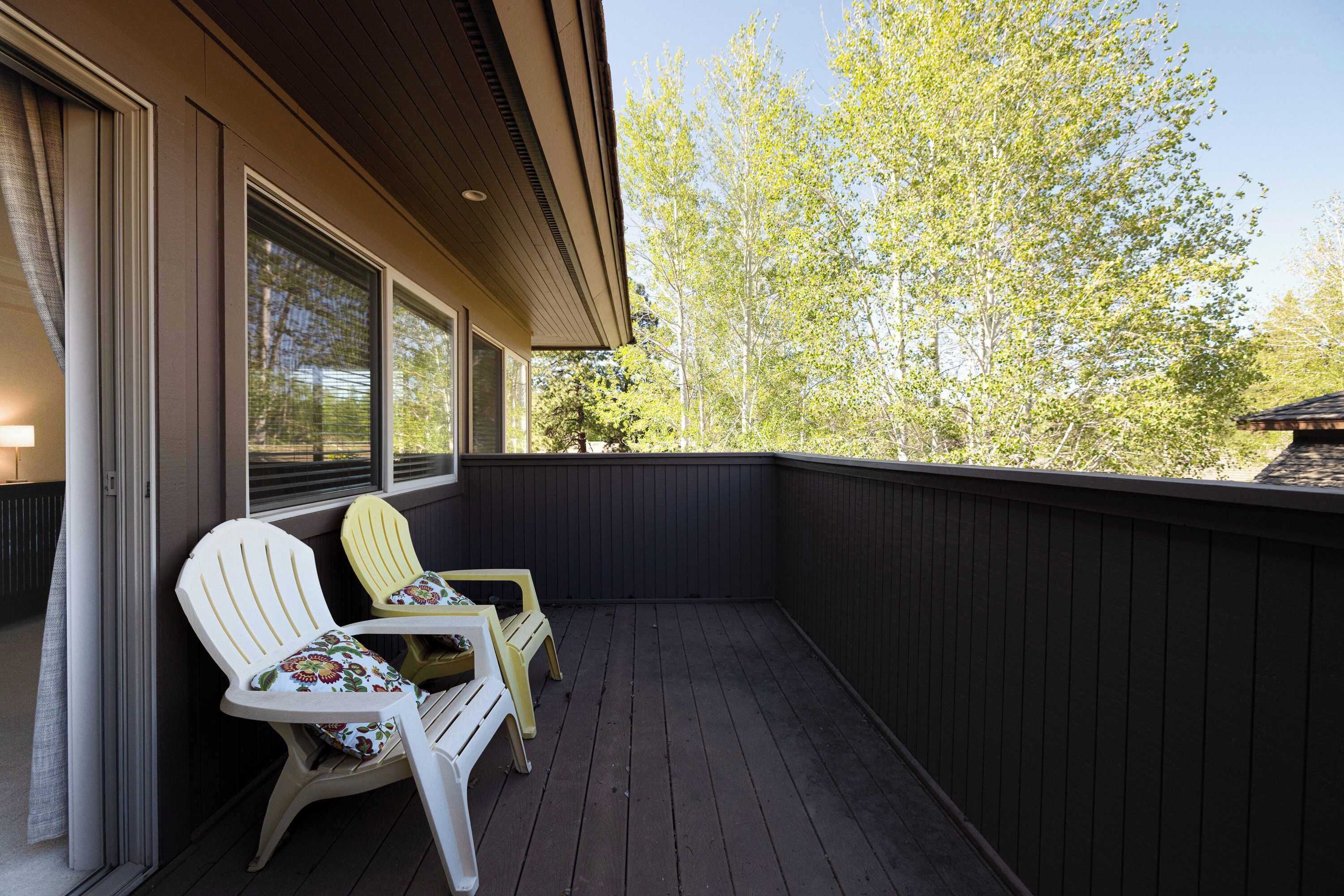 A covered outdoor area with two Adirondack chairs and a table, wooden deck, and a view of trees.