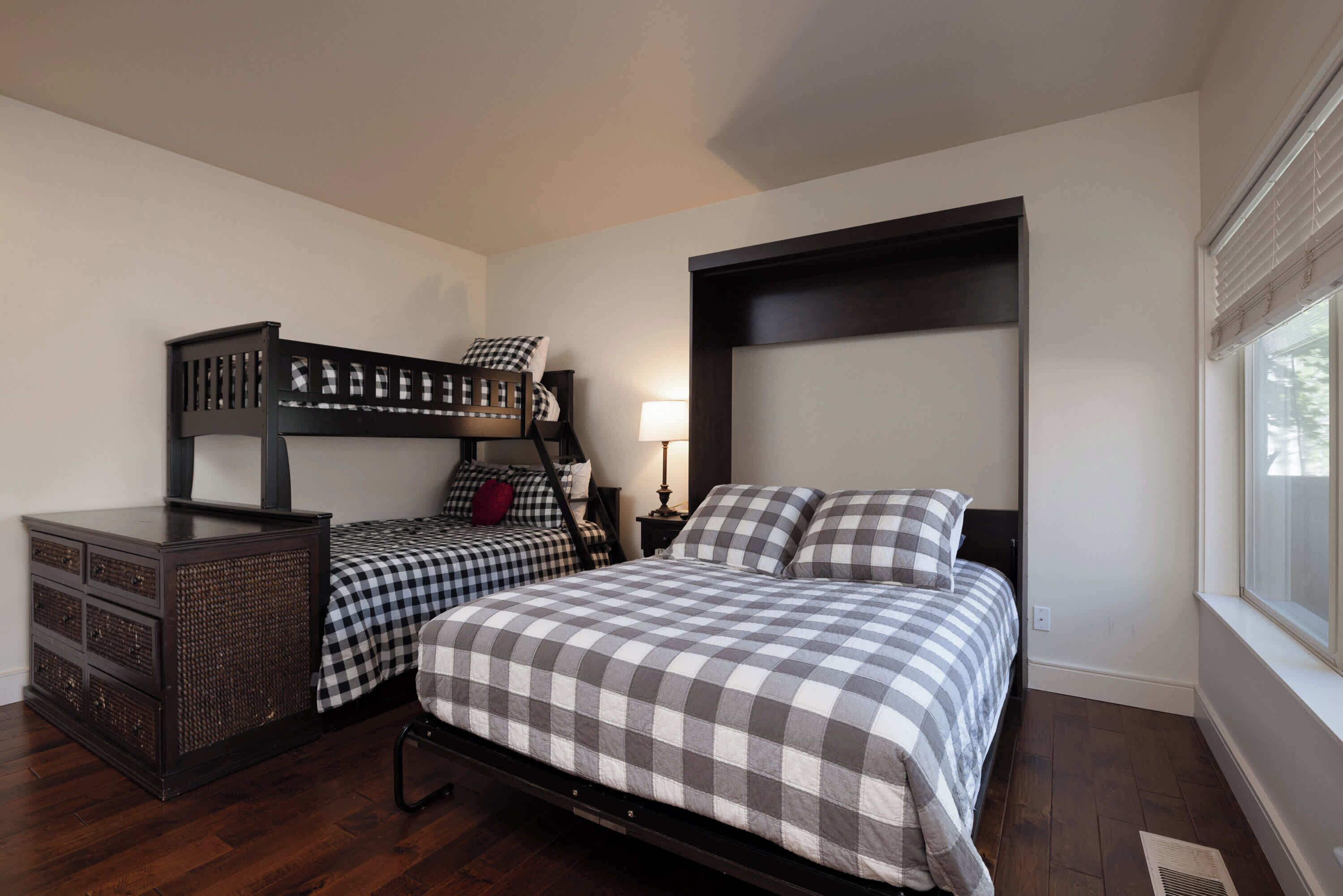 A bedroom with a bunk bed, a dresser, a bedside table with a lamp, and a window with blinds.