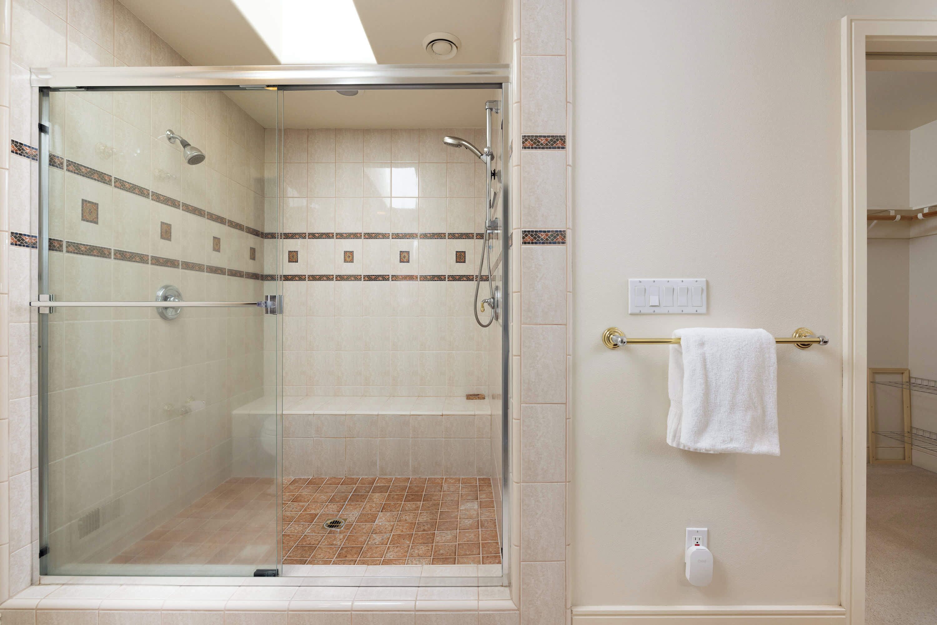 A glass-enclosed shower with a tiled floor, a towel rack with a white towel, and a wall-mounted electrical outlet.