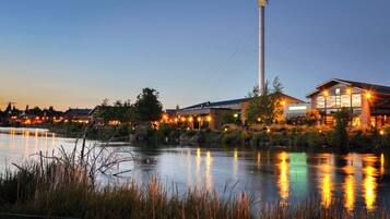 A riverside hotel with a prominent tower, reflecting lights on the water, surrounded by greenery and other buildings.