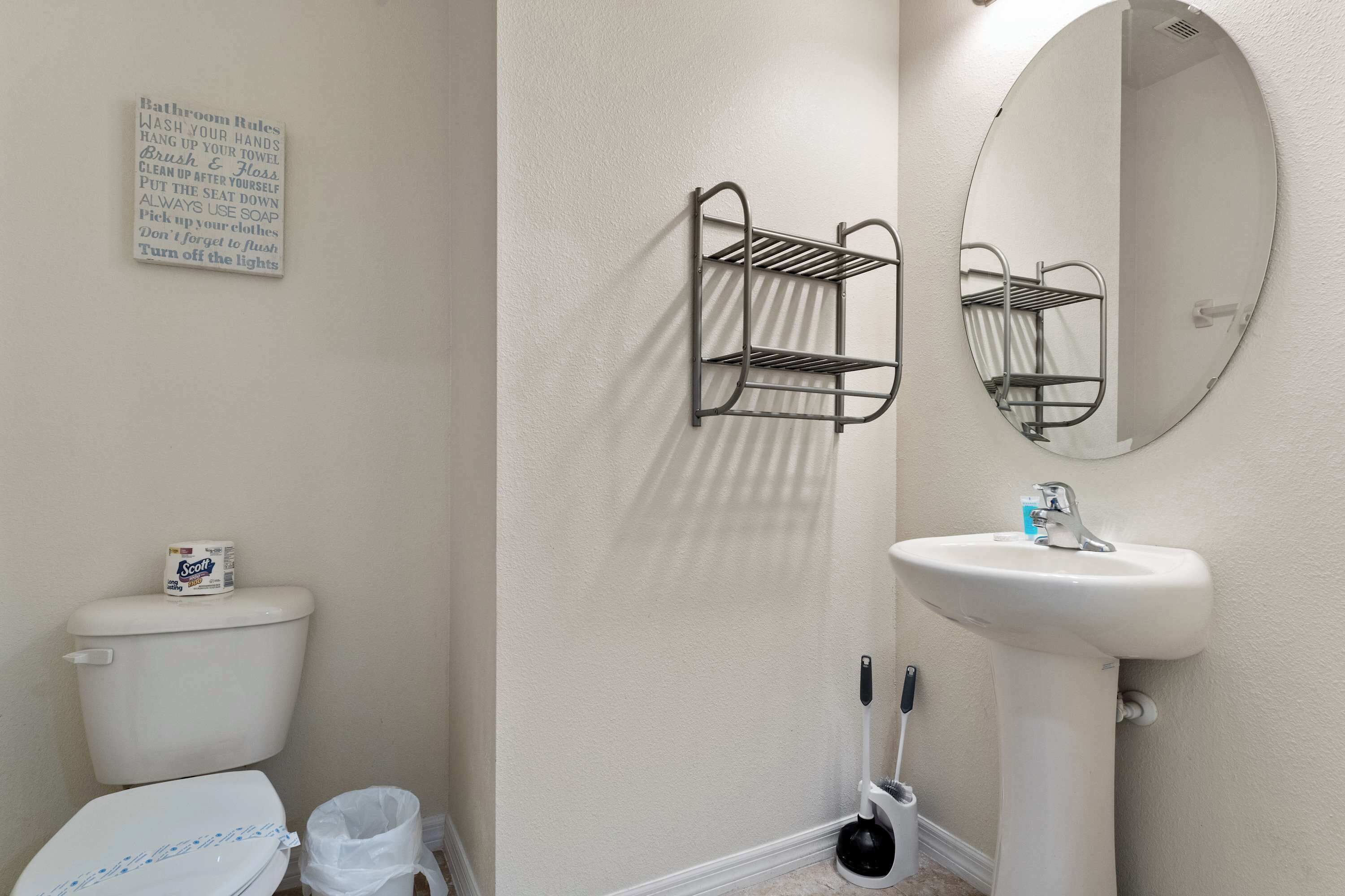 Un baño con inodoro, un lavamanos blanco, un espejo, una repisa metálica y un cartel con normas del baño.