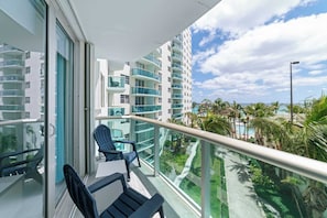 A balcony with a glass railing, a chair, and a view of a multi-story building with balconies and a pool area. - Ocean Wind Luxury Beachfront Condo With Pool & Gym (Hollywood)