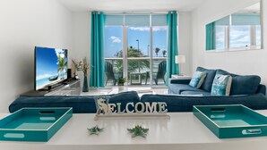 Ein modernes Wohnzimmer mit großem Fernseher, lindgrünen Vorhängen, einer Couch und einem Schild mit der Aufschrift 'WELCOME'.