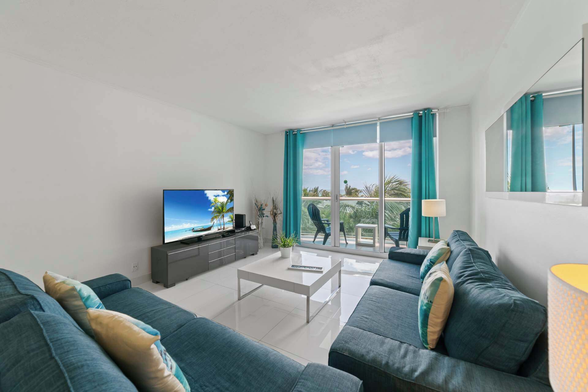 A modern living room with a flat-screen TV, a sofa set, a coffee table, and a balcony with a view of palm trees.