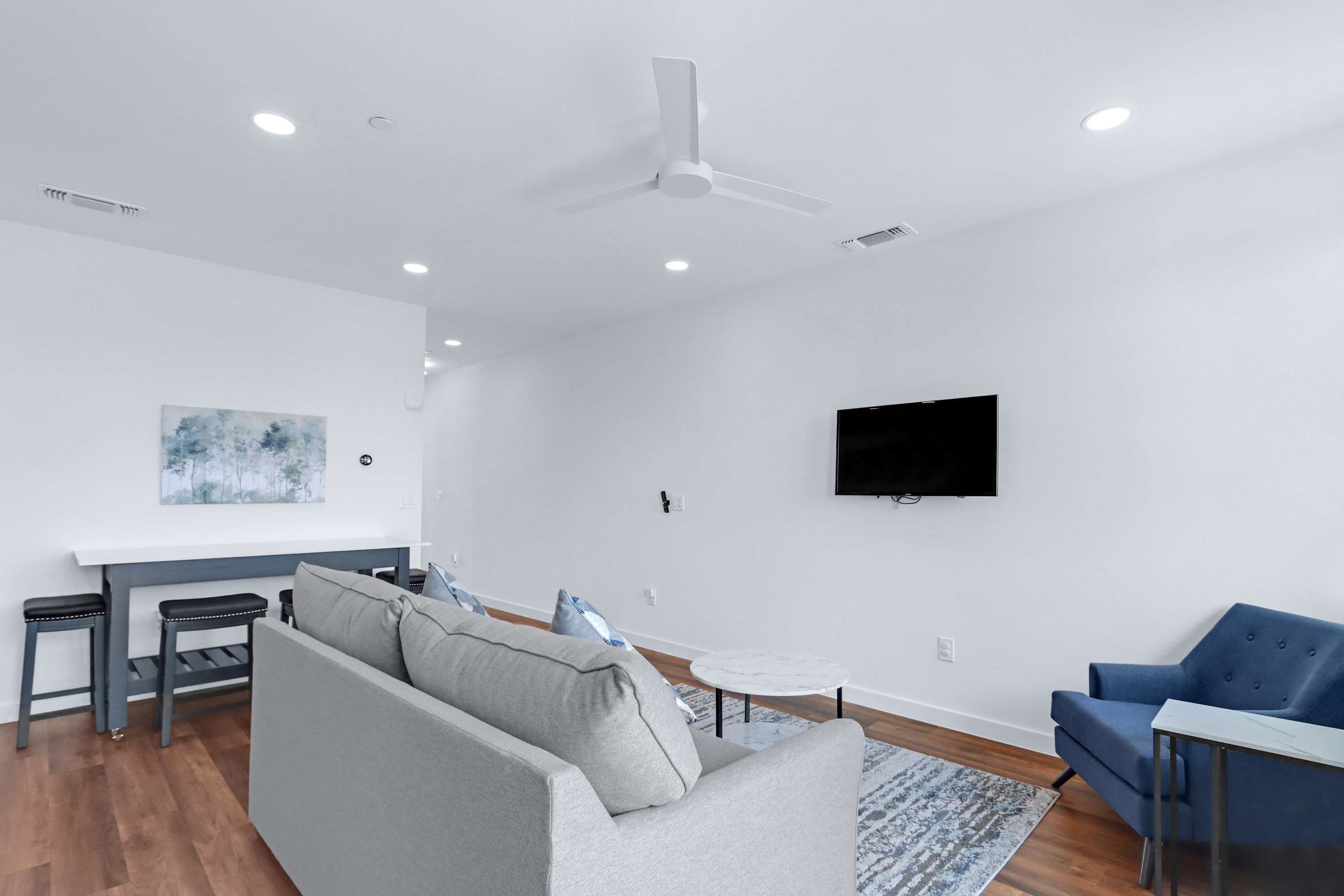 Una sala de estar moderna con un sofá gris, un sillón azul, una mesita redonda y un televisor de pantalla plana montado en la pared.