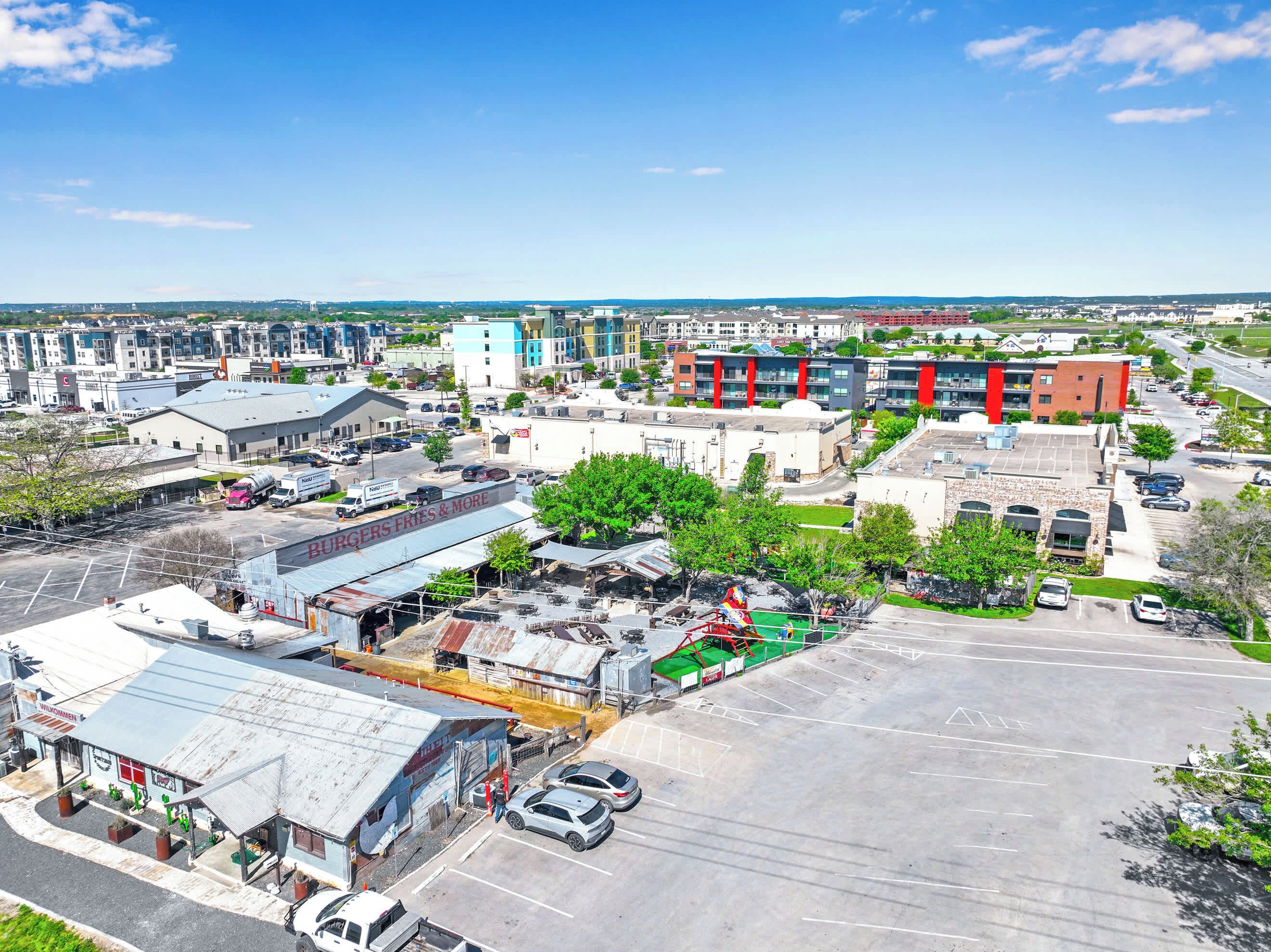 Vista aérea de una zona comercial con varios edificios, incluyendo un estacionamiento con coches y un amplio espacio abierto.