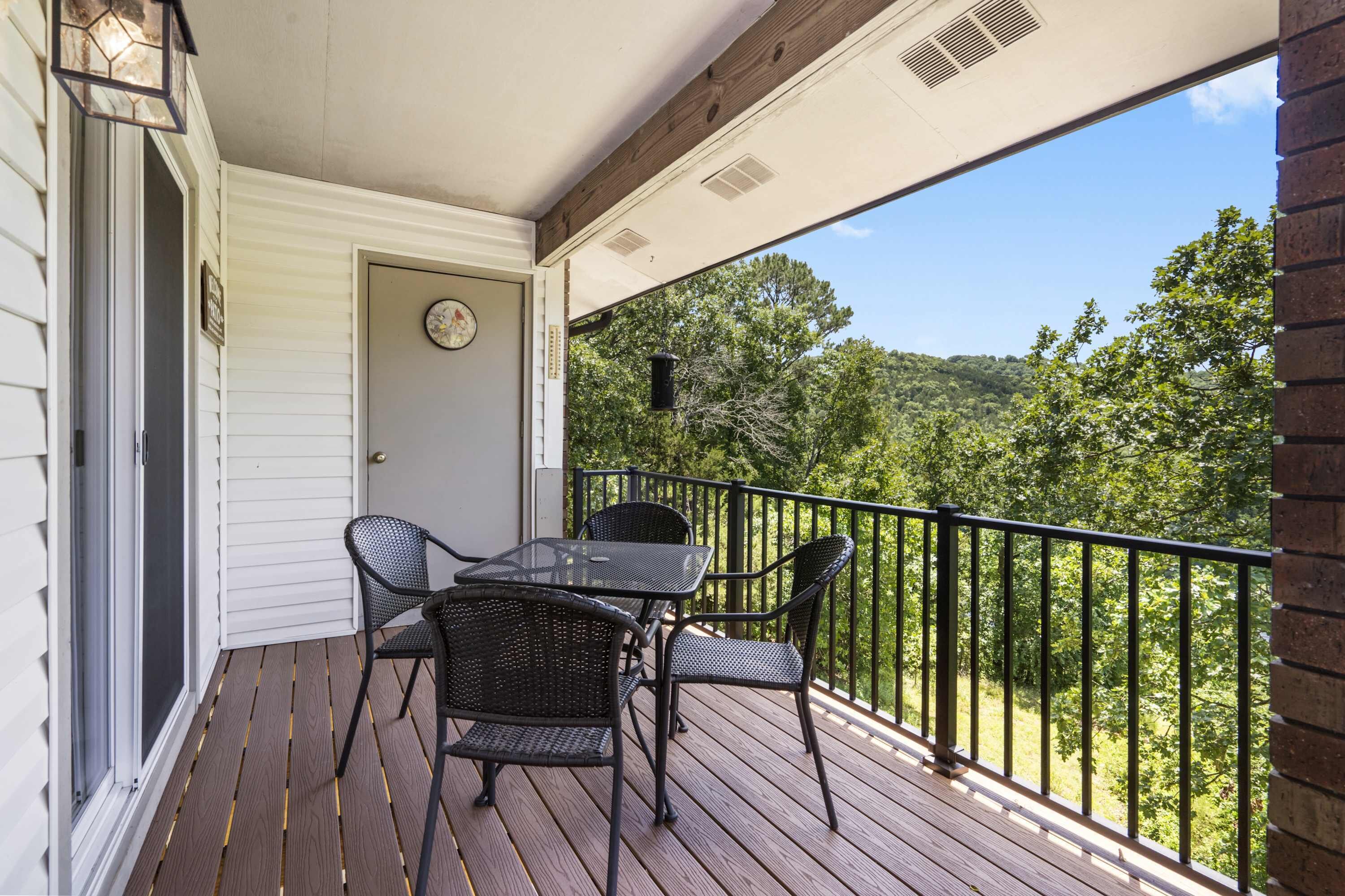 A wooden deck with a table and chairs, a door with a clock, and a view of trees and hills.