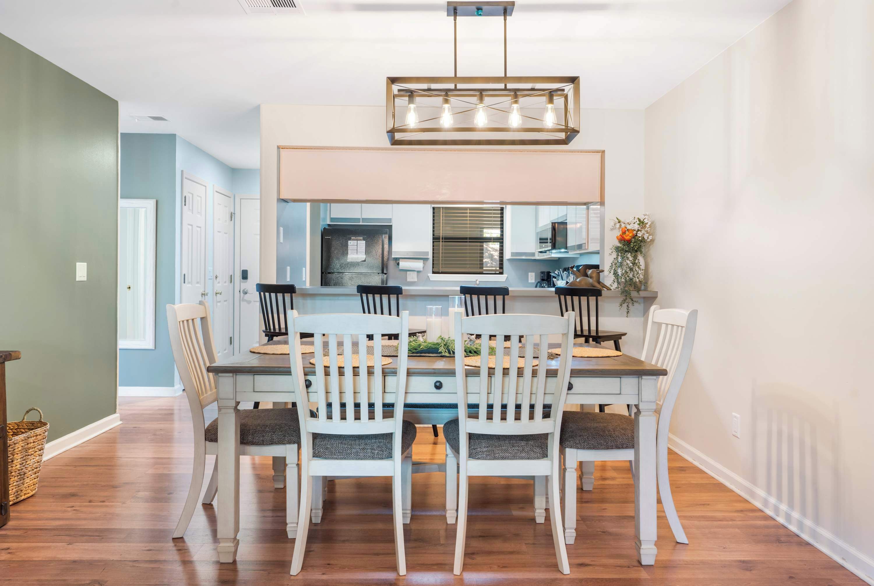 Un comedor con una mesa de madera y sillas blancas, una lámpara de araña moderna, y vista a la cocina.