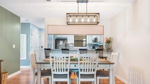A dining area with a wooden table and white chairs, a modern chandelier, and a view into the kitchen. - My Kind of Getaway - Chic 2 Bdr! (Branson)
