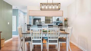 Un comedor con una mesa de madera y sillas blancas, una lámpara de araña moderna, y vista a la cocina.