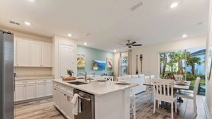 A modern kitchen with white cabinetry, a central island, and a dining area with white chairs. - Mint to Be (Kissimmee)