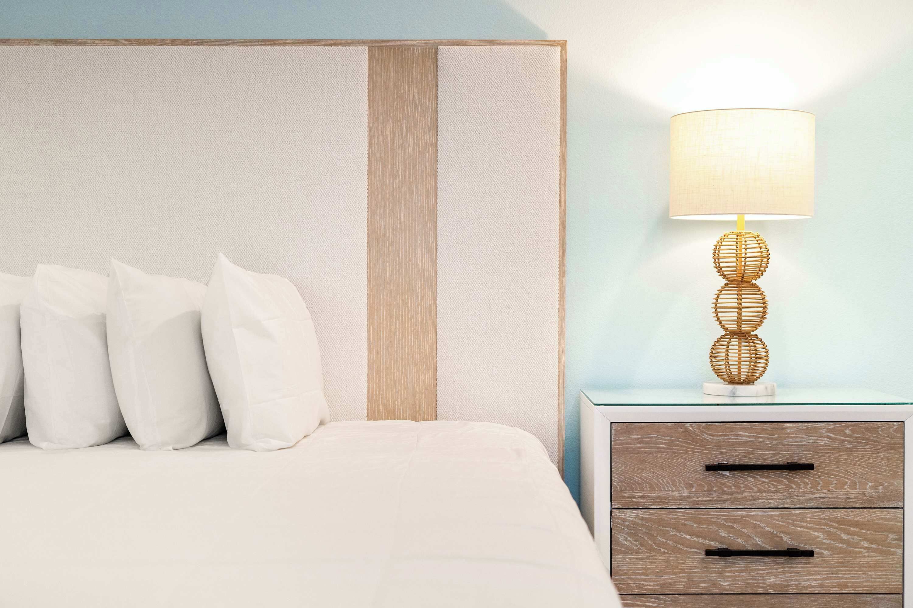 A neatly made bed with white bedding and pillows, a bedside table with a lamp, and a wall with a textured panel.