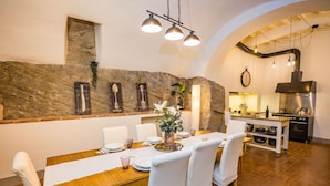 A dining area with a stone wall, a wooden table set for four, white chairs, and a kitchen in the background.