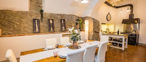 Un comedor con pared de piedra, una mesa de madera dispuesta para cuatro, sillas blancas y una cocina al fondo.