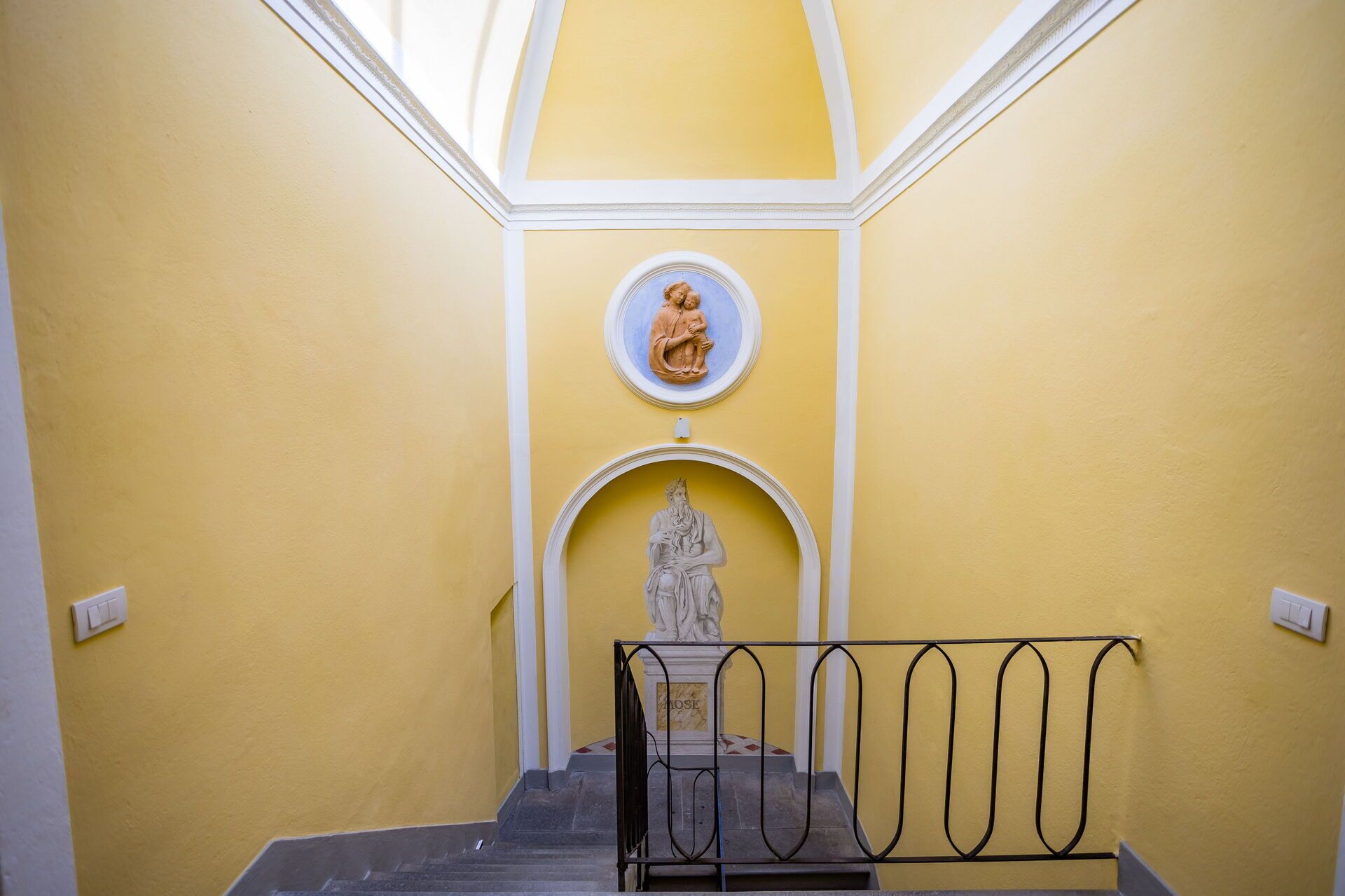 A staircase with a statue at the bottom, a circular plaque above, and a metal railing in the foreground.