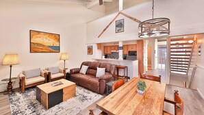 A spacious living area with a brown leather sofa, two armchairs, a wooden coffee table, and a dining table with chairs. A kitchen is visible in the background with wooden cabinets and a hanging light fixture.